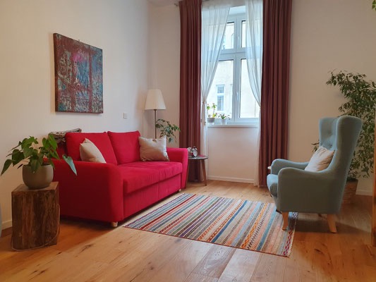 Praxisraum mit rotem Sofa, hellblauem Sessel, Holzboden und Pflanzen